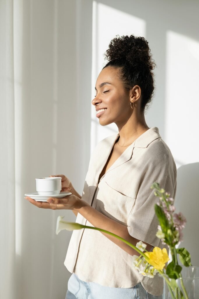 A woman stands smiling with a cup and saucer in natural light, creating a serene morning atmosphere.